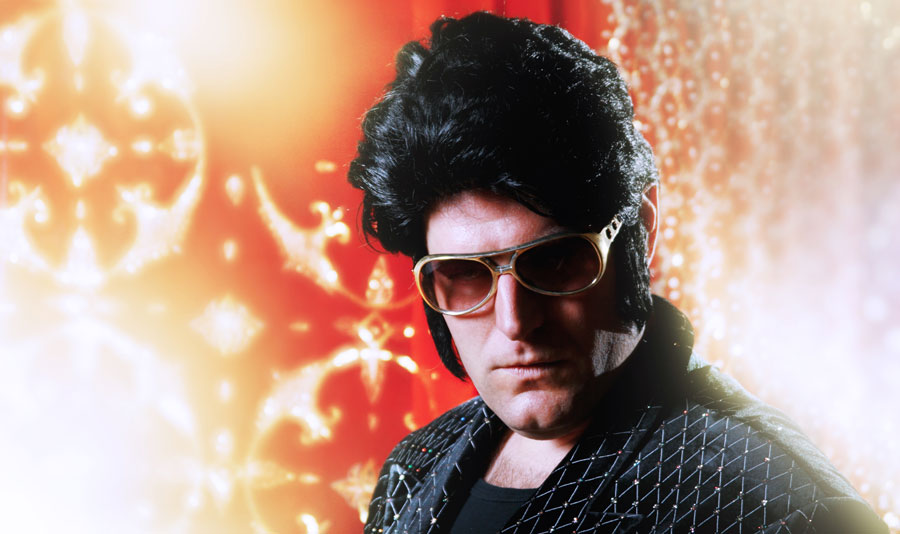 Elvis impersonator with pompadour wig, sunglasses, and a rhinestone-studded jacket against a vibrant, warm background.